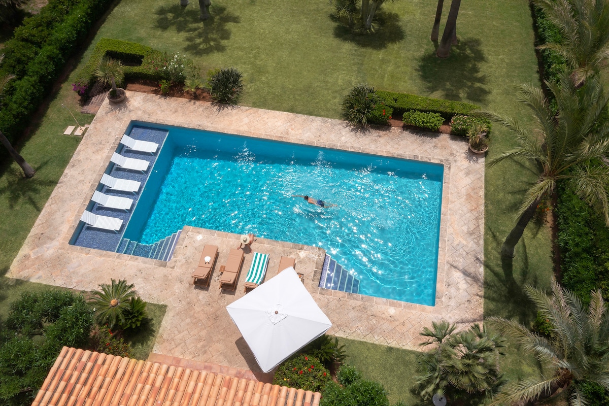 An aerial view of a large swimming pool surrounded by lush greenery. Sun loungers are arranged along the pool's edge, and a shaded area with a parasol and chairs is visible. The clear water reflects the sky, inviting relaxation and leisure.