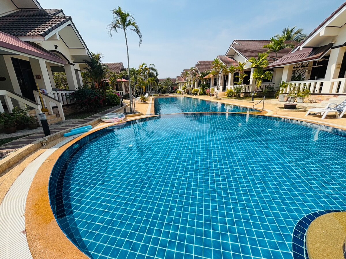 A spacious swimming pool is surrounded by well-maintained tropical landscaping. Multiple lounge chairs are positioned along the poolside. The clear blue water is inviting and reflects the sunny sky, while the surrounding residences are visible in the background.