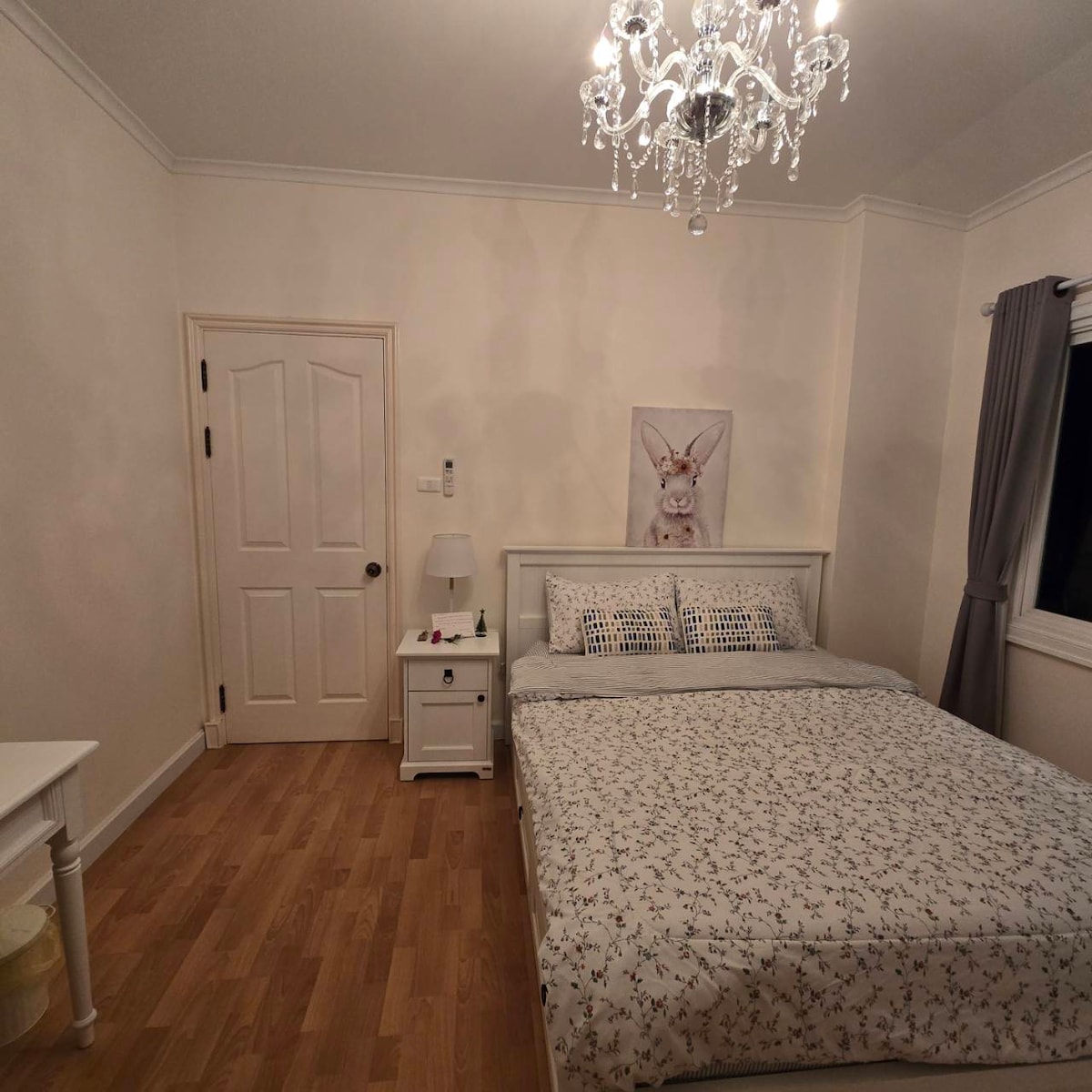 A serene bedroom features a spacious bed adorned with a patterned blanket. A small nightstand with a lamp is positioned beside the bed. Natural light enters through a dark-curtained window, illuminating a delicate chandelier overhead and a framed art piece on the wall.