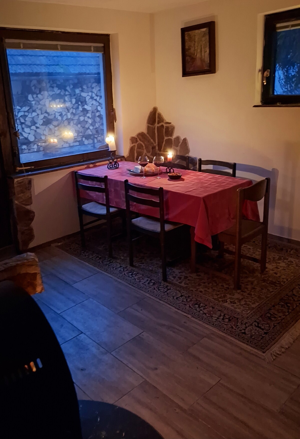 An intimate dining area is presented with a pink tablecloth covering a wooden table surrounded by six chairs. Soft candlelight flickers on the table, adding warmth. A stone wall is visible in one corner, while a large window partially obscured by shades offers glimpses of the outside.