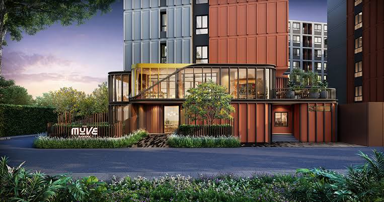 The modern building exterior is highlighted by a mix of angular design elements and large glass windows. Lush greenery surrounds the entrance, which features a welcoming sign. The facade combines various color panels, creating a contemporary and vibrant appearance against a tranquil sunset backdrop.