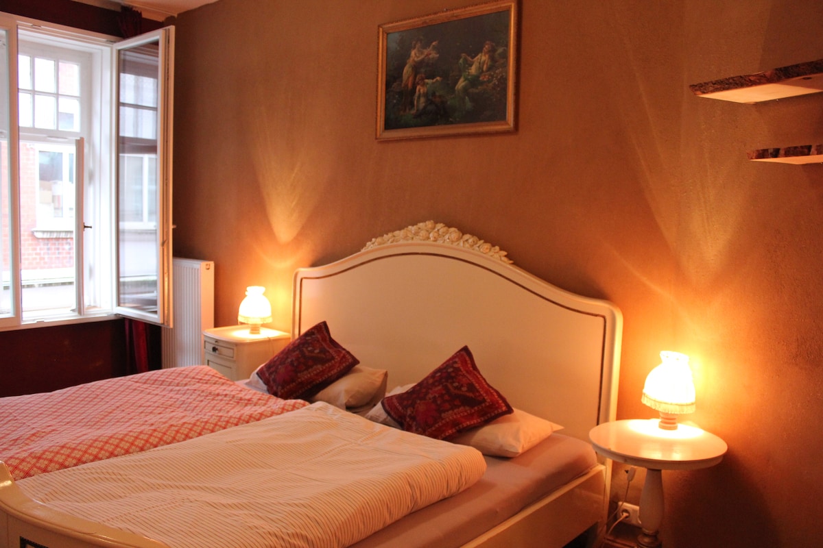 A spacious bed is centered in the room, adorned with patterned cushions and a light-colored duvet. Soft lamps on either side provide gentle illumination. A piece of wall art depicting a serene landscape hangs above the bed, and a window with partial curtains allows natural light to filter in.