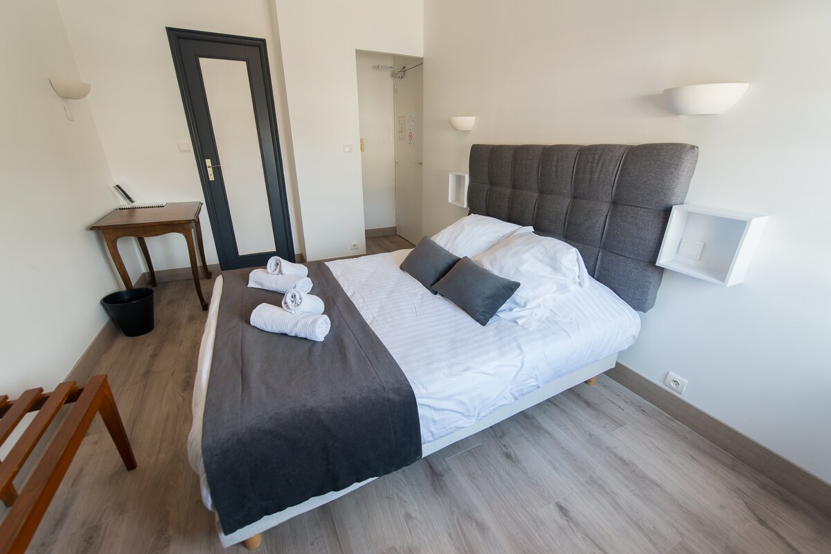 The room features a spacious bed with crisp white linens and gray accents, complemented by two decorative pillows. A wooden table is positioned against the wall, alongside a wastebasket. Light from the window enhances the minimalist decor, while the warm wooden flooring adds a subtle touch.
