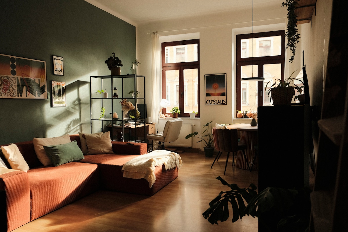 The living room features warm lighting, highlighting a spacious, sectional sofa in earthy tones. A bookshelf filled with plants and decor items is positioned against a green accent wall. Large windows allow natural light to fill the space, creating a cozy atmosphere for relaxation.