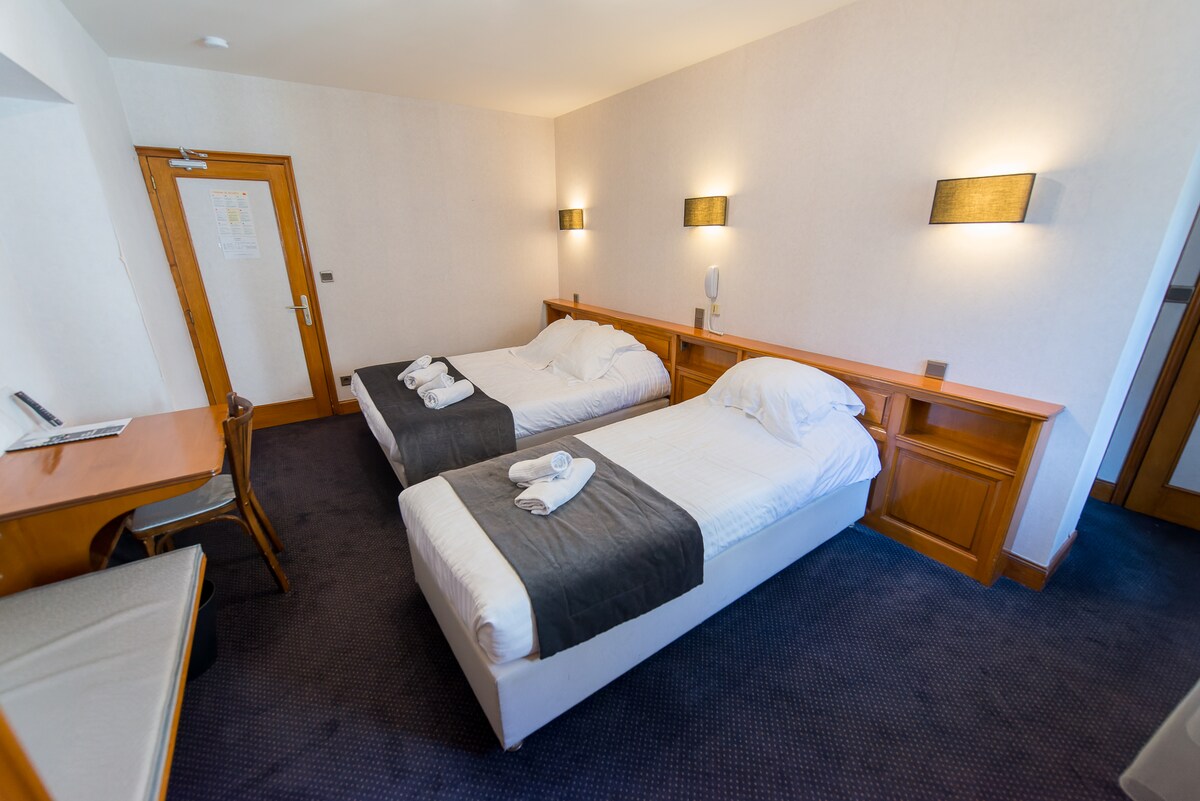 A well-lit guest room featuring two single beds adorned with white linens and dark grey throws. A wooden desk and chair are present, alongside a large wall-mounted lamp. Soft carpeting covers the floor, creating a comfortable environment, while a door leads to another section of the space.