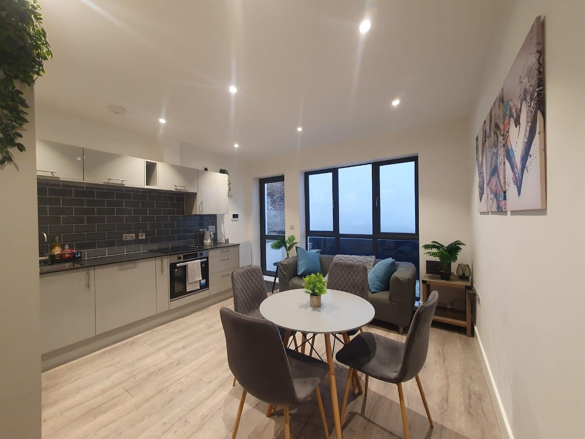 An open-plan living area features modern kitchen cabinets and a sleek countertop. A round dining table is surrounded by four grey chairs. Natural light enters through large windows, showcasing a tranquil indoor space complemented by decorative plants and artwork on the walls.