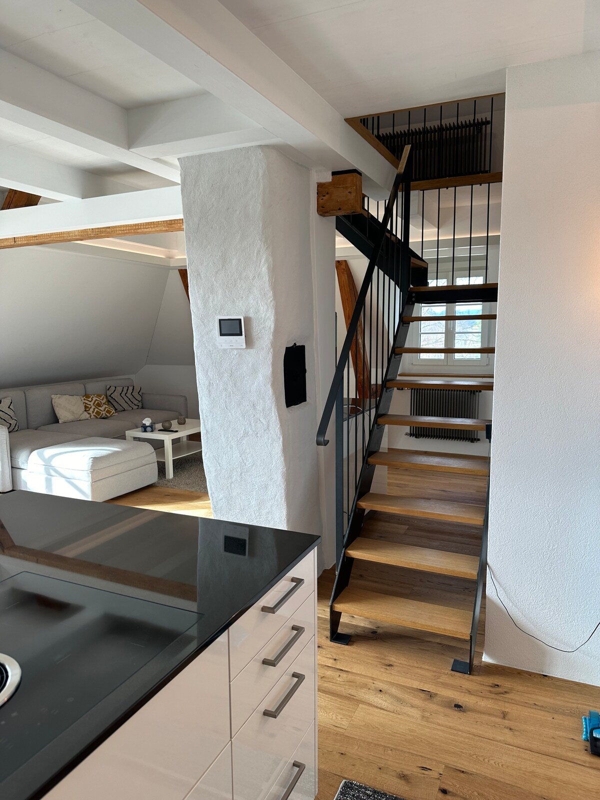 A staircase with a metal railing ascends to a loft area, surrounded by light wood beams. The open layout reveals a cozy seating arrangement with a light-colored couch and a table, enhancing the inviting atmosphere of the living space.