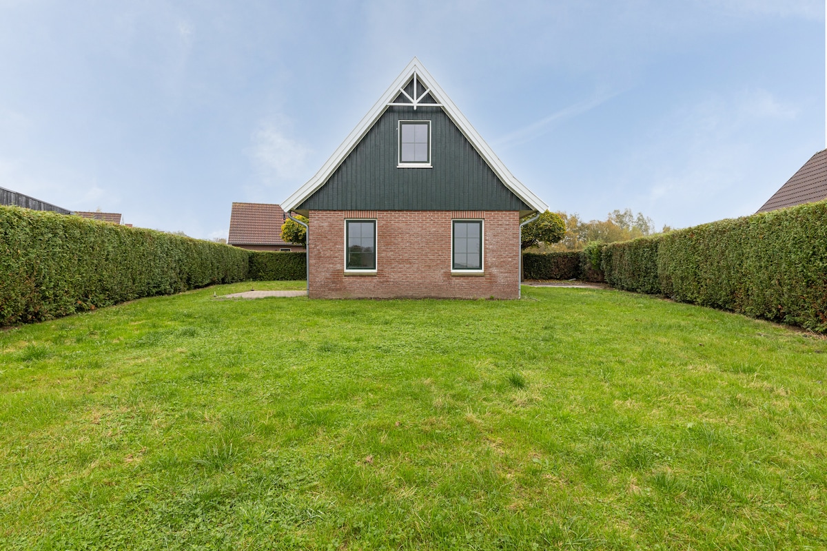 The exterior of the house displays a modern design with a dark green upper section and a brick lower section. Large windows invite natural light, while a neatly maintained lawn is surrounded by hedges, providing a sense of privacy and openness.