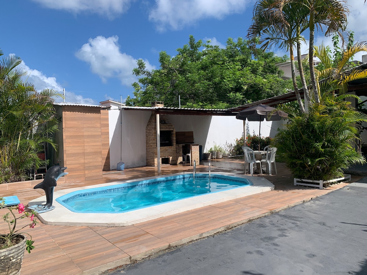 A private outdoor space features a rectangular swimming pool surrounded by wooden decking and tropical plants. A barbecue area is visible against a neutral wall, along with multiple chairs and an umbrella for shade. The sky is clear, allowing natural light to illuminate the area.
