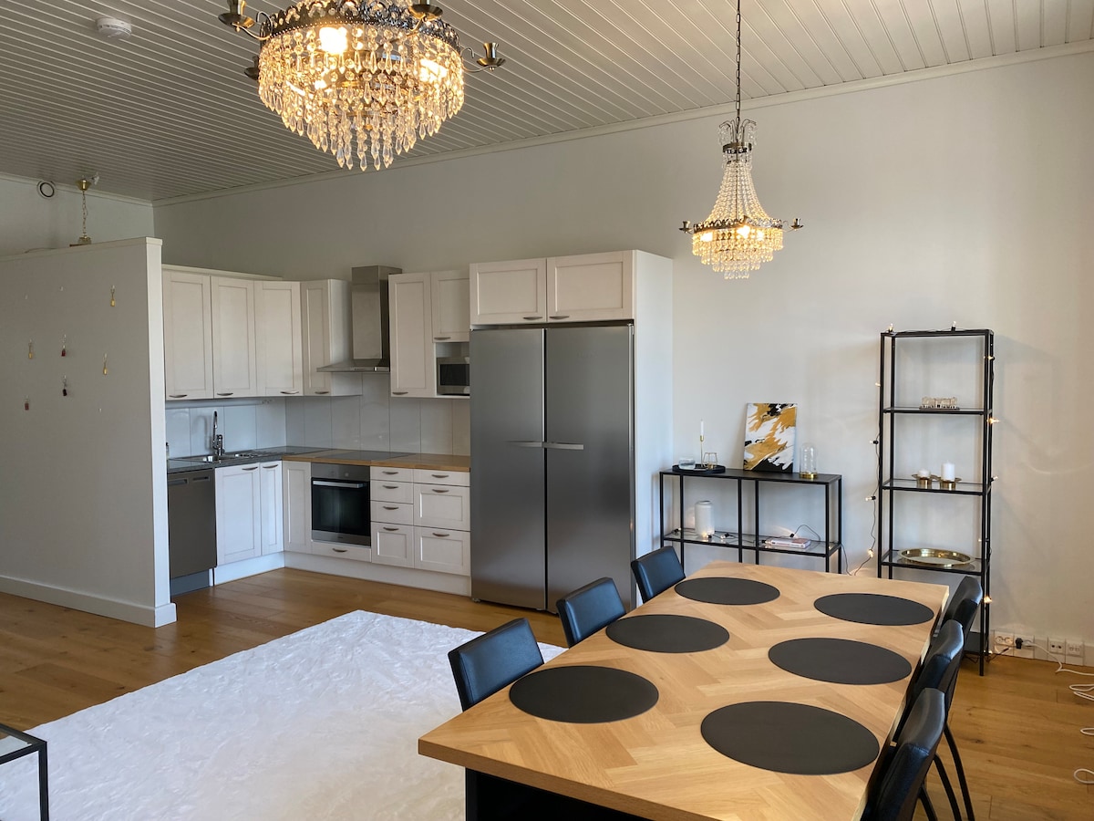 A modern open kitchen and dining area is displayed, featuring a spacious wooden table surrounded by black chairs. Bright light fixtures hang from the ceiling, illuminating the stylish kitchen equipped with white cabinetry and stainless steel appliances. A soft rug lies beneath the table, enhancing the overall design.