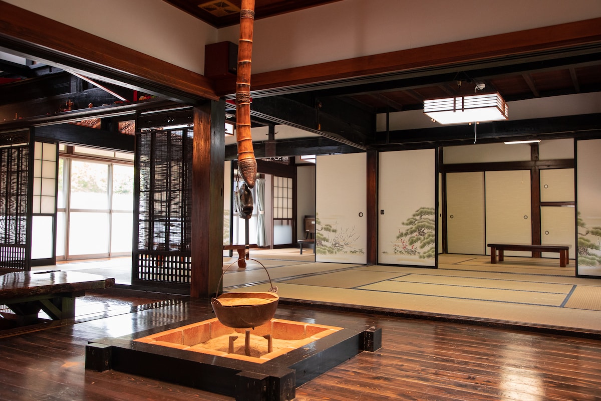 A traditional interior is showcased with polished wooden floors and open spaces. A decorative Irori fireplace is centered in the room, surrounded by tatami mats and sliding shoji doors that lead to bright, airy areas filled with natural light.