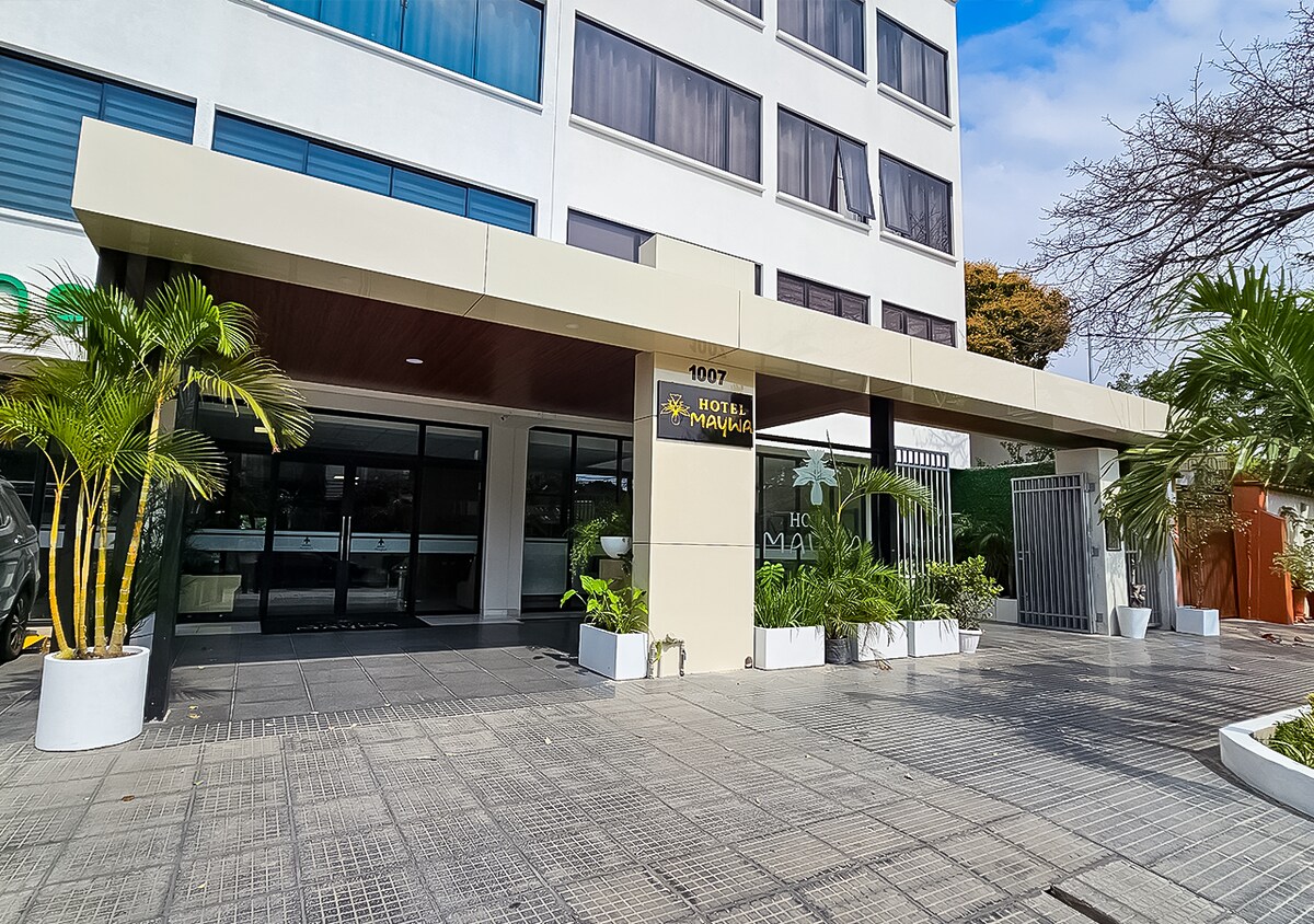 The entrance of Hotel Maywa features a modern façade with a large canopy. Potted plants frame the entryway, and the glass doors provide a clear view into the lobby. The exterior is complemented by a well-maintained walkway and decorative landscaping.