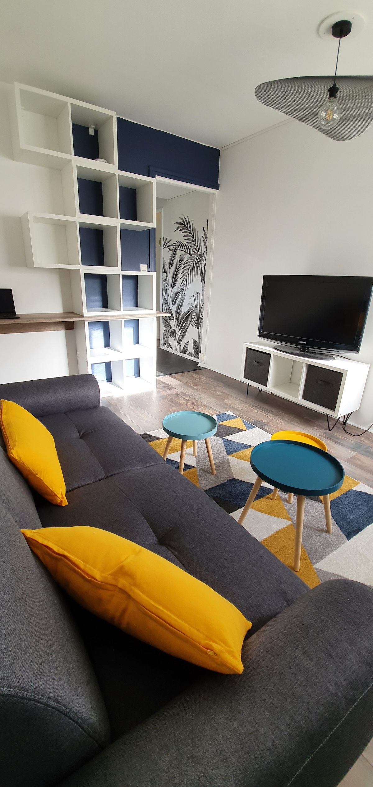 A modern living area features a dark grey sofa adorned with yellow cushions, complemented by two small round tables. A television stands on a white media console against a patterned accent wall, while a bookshelf adds visual interest to the space.