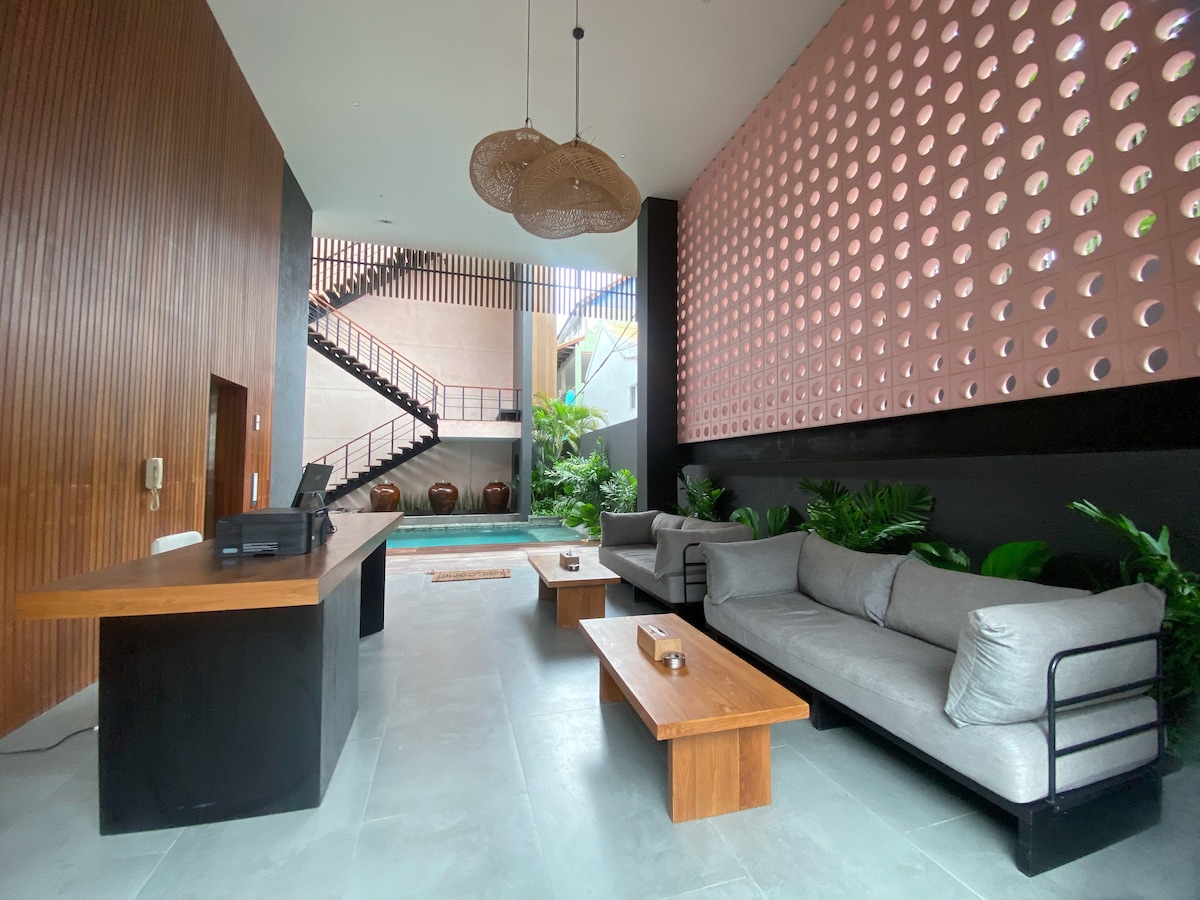 A modern reception area is highlighted by a wooden front desk, with soft gray couches arranged around a wooden coffee table. Natural light filters through circular cutouts in the wall, illuminating lush greenery and a glimpse of a pool in the background.