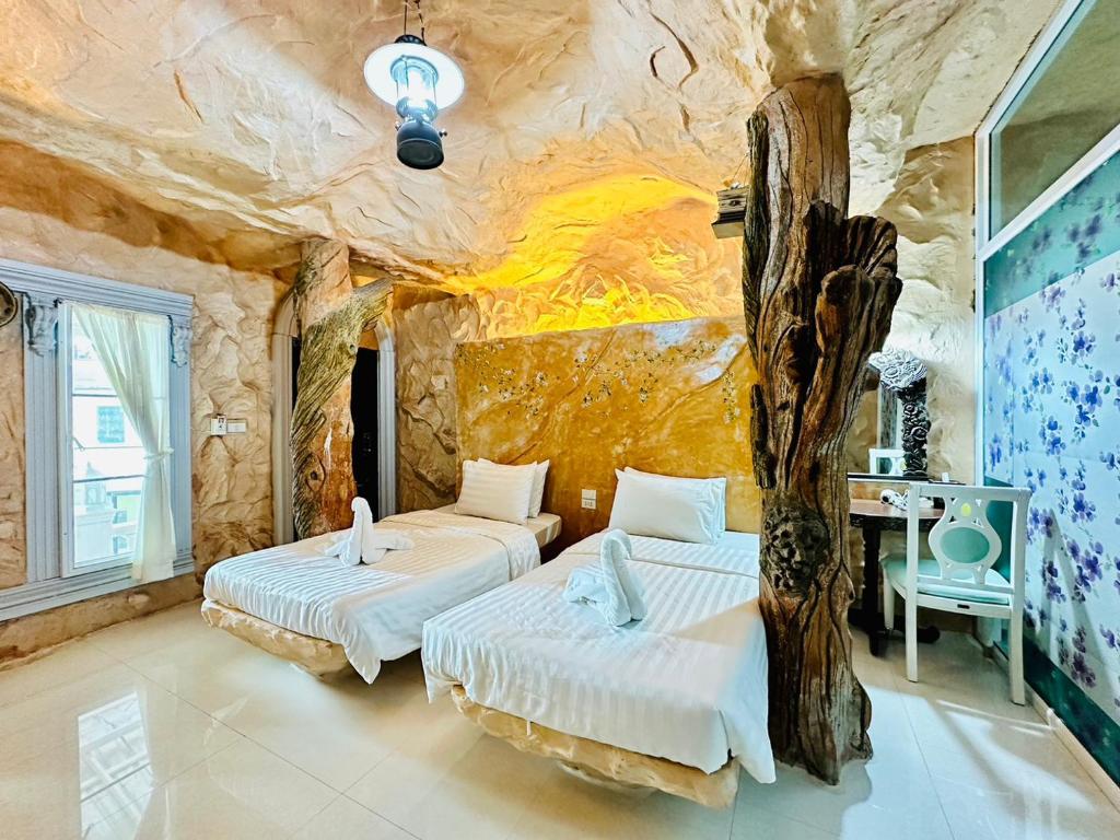 A unique accommodation features textured stone walls that mimic a natural cave setting. Two beds with crisp white linens are positioned on a polished floor. Decorative elements, including wooden accents, add character, while a small table is visible against a colorful mural of flowers.