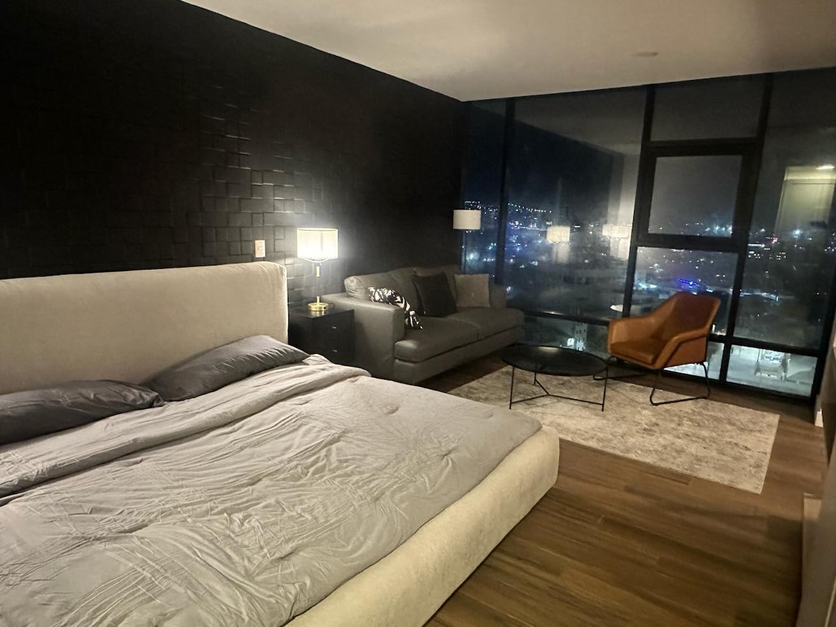 An inviting bedroom features a spacious bed with neutral bedding and a textured headboard. A dark sofa and a contemporary armchair are positioned near a sleek coffee table, offering comfort. Large windows provide a view of the city skyline, softly illuminated at night.
