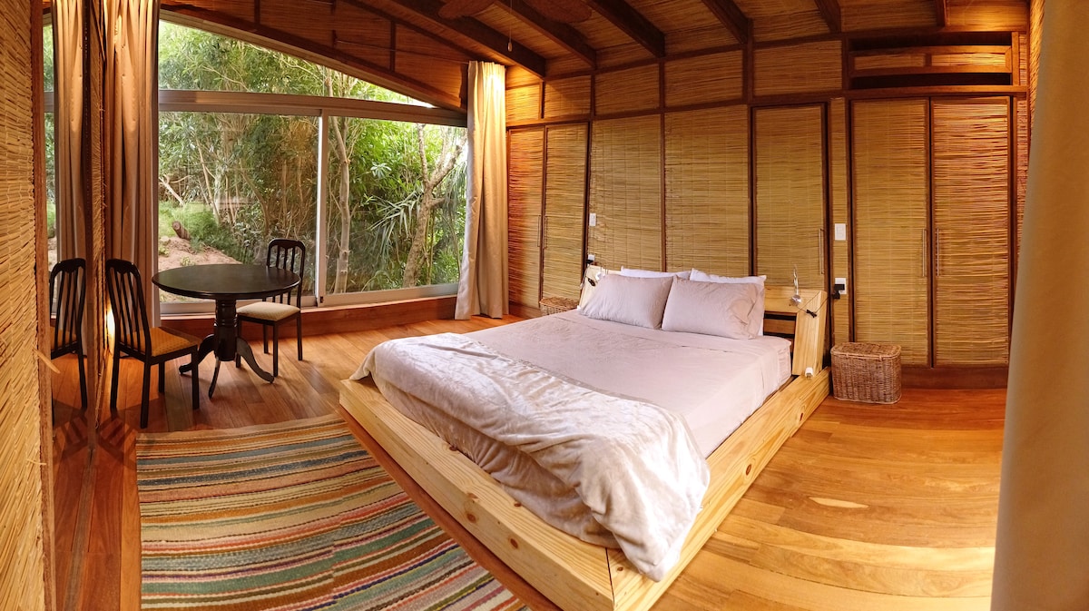A spacious room features a wooden platform bed with soft bedding. A round dining table and chairs are set against a backdrop of large windows, revealing lush greenery outside. Natural wood accents and bamboo walls create a warm atmosphere.