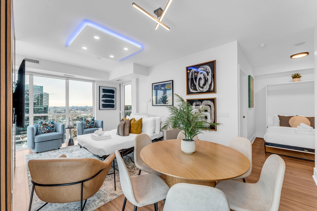 A modern living space showcases a round wooden dining table surrounded by sleek chairs. Large windows reveal expansive city and lake views, while a cozy seating area features a plush couch. Accent lighting enhances the contemporary design of the room.
