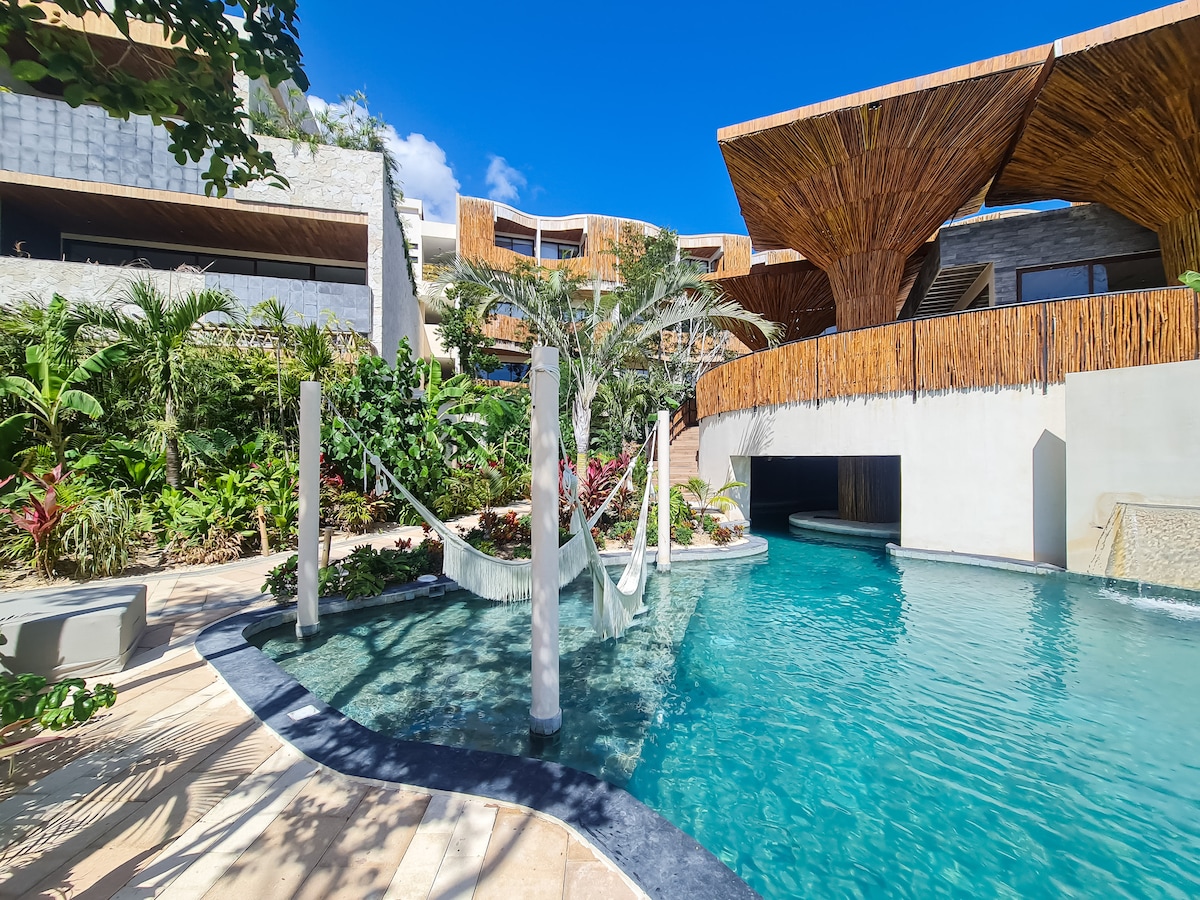 A serene pool area is surrounded by lush tropical plants and features wooden structures with thatched roofs. A hammock is suspended gracefully over the water, inviting relaxation under the clear blue sky. The design allows for seamless integration with the natural landscape.