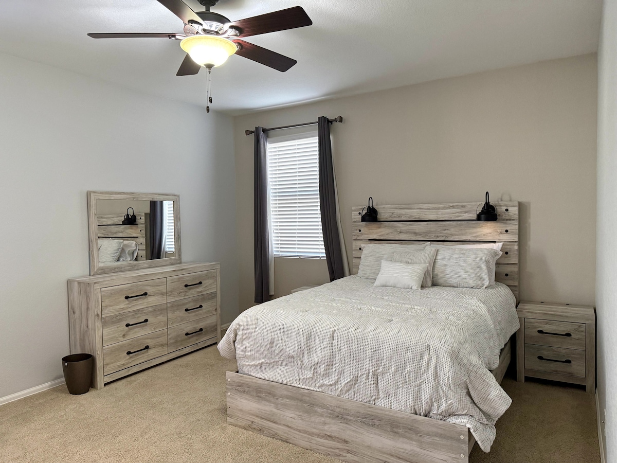 A well-appointed bedroom features a spacious bed with a light grey quilt, flanked by two bedside tables. A matching dresser and mirror complement the decor. Natural light enters through a window adorned with dark curtains, while a ceiling fan adds comfort.