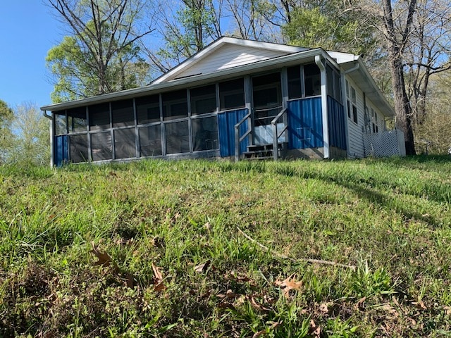 A single-story cottage is set on a grassy incline, featuring a screened-in porch with a blue accent. Surrounding trees offer a natural backdrop, while the structure is accessed by a set of stairs leading to the porch. The peaceful environment is highlighted by a clear sky.