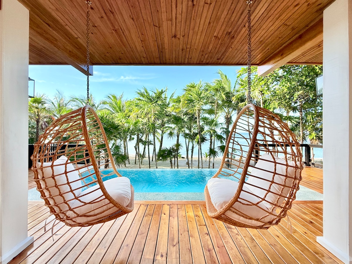 Two cozy hanging swing chairs are positioned on a wooden deck, facing a private infinity pool. Lush palm trees are visible in the background, with the ocean beyond, creating a serene and inviting outdoor space.