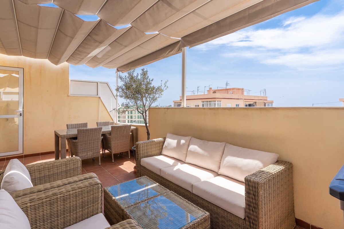 A spacious terrace is furnished with a sofa and a glass-top coffee table, surrounded by a shaded canopy. A dining table with wicker chairs is also present. A view of the sky and other buildings can be seen in the background, creating a relaxed outdoor setting.
