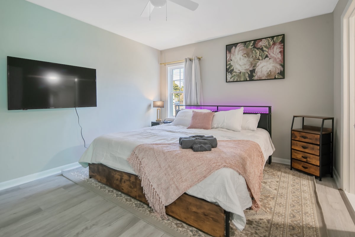 A spacious bedroom features a large king bed with a light-colored bedspread and decorative throw. A mounted flat-screen TV is positioned on the wall opposite the bed. Natural light enters through a window, and gentle tones of grey are complemented by a floral wall art piece.