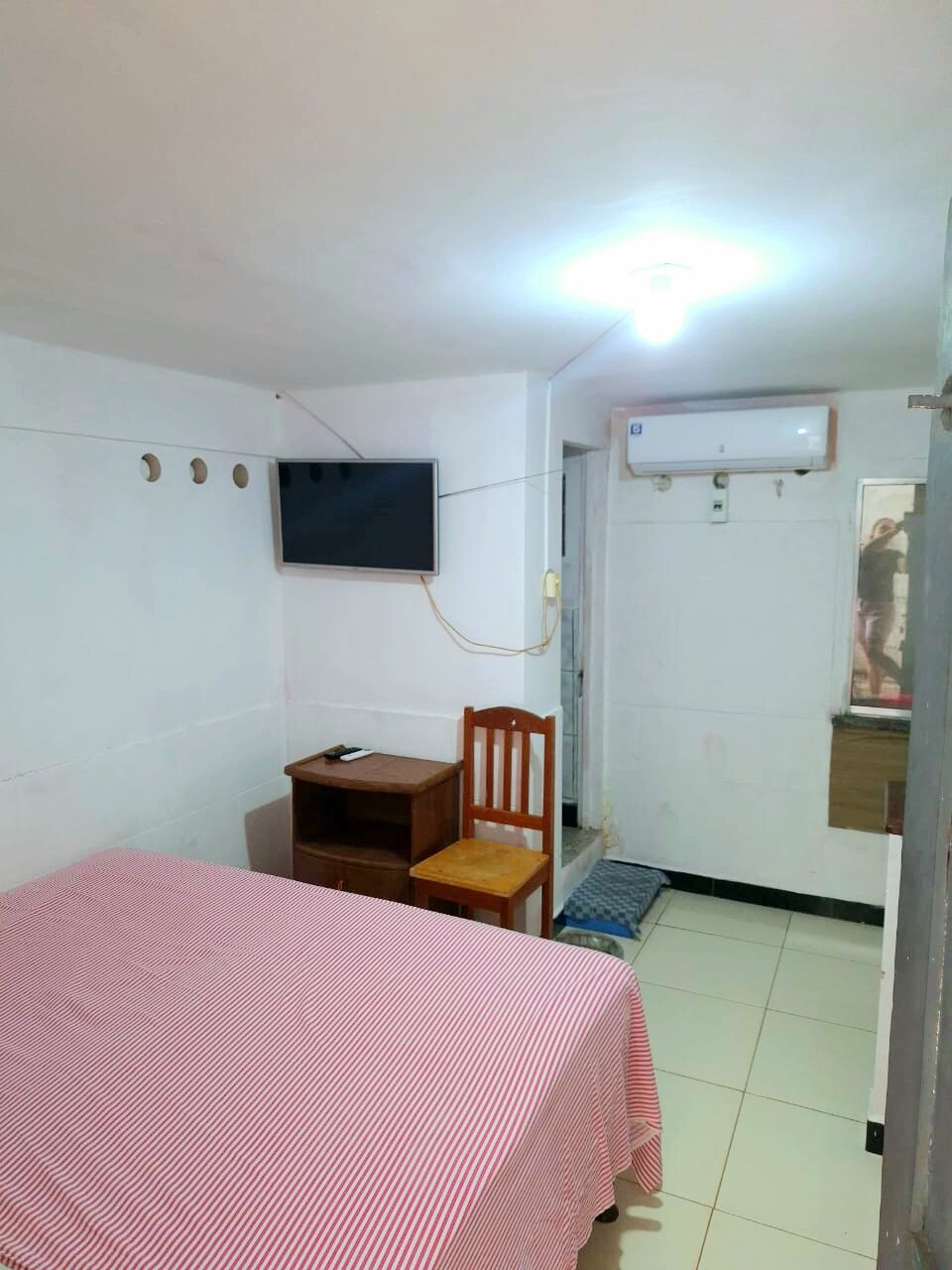 A simple room is presented with a red and white striped bedspread covering a spacious bed. A wooden chair and small desk sit adjacent to the bed. The wall-mounted TV and air conditioning unit contribute to the room’s comfort. Light is provided by a ceiling fixture.
