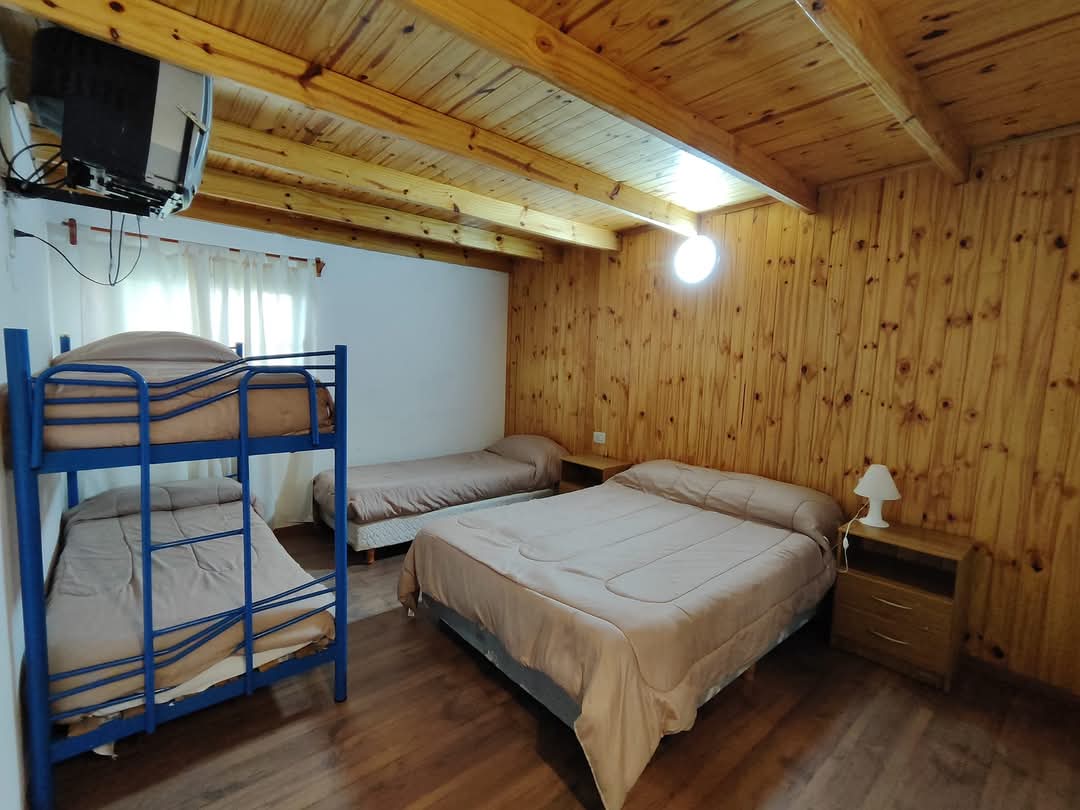 A room features wooden walls and a ceiling with exposed beams. A spacious double bed is positioned alongside a single bed and a blue bunk bed. A bedside table with a lamp is present next to the double bed. Soft light enters through a partially draped window.