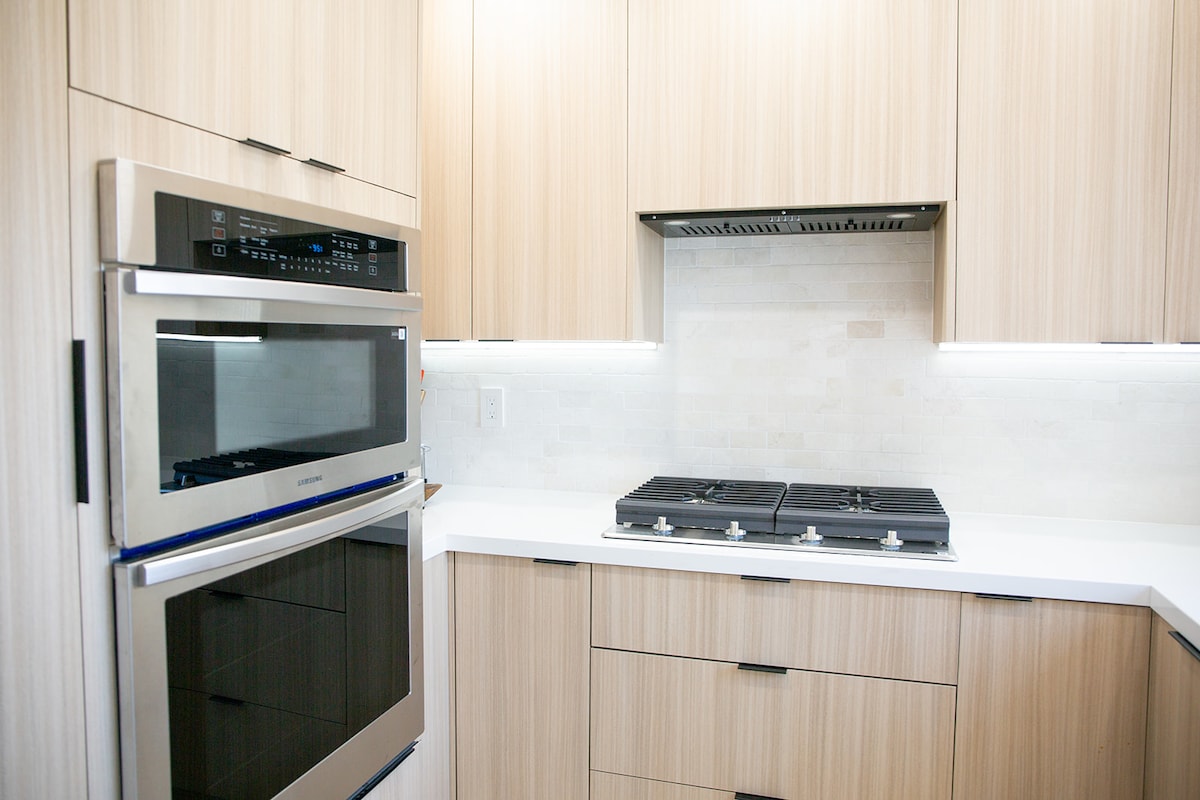 A modern kitchen showcases light wood cabinetry with sleek lines. Stainless steel appliances, including an oven and stovetop, are featured. A white countertop extends against a neutral tiled backsplash, while under-cabinet lighting provides a subtle illumination.
