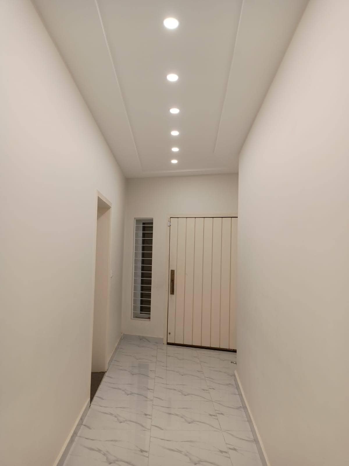 A well-lit hallway features recessed ceiling lights, providing even illumination along the smooth marble floor. The walls are painted in a neutral color, enhancing the space's openness. A door leads to the outside, while a window adds natural light to the corridor.