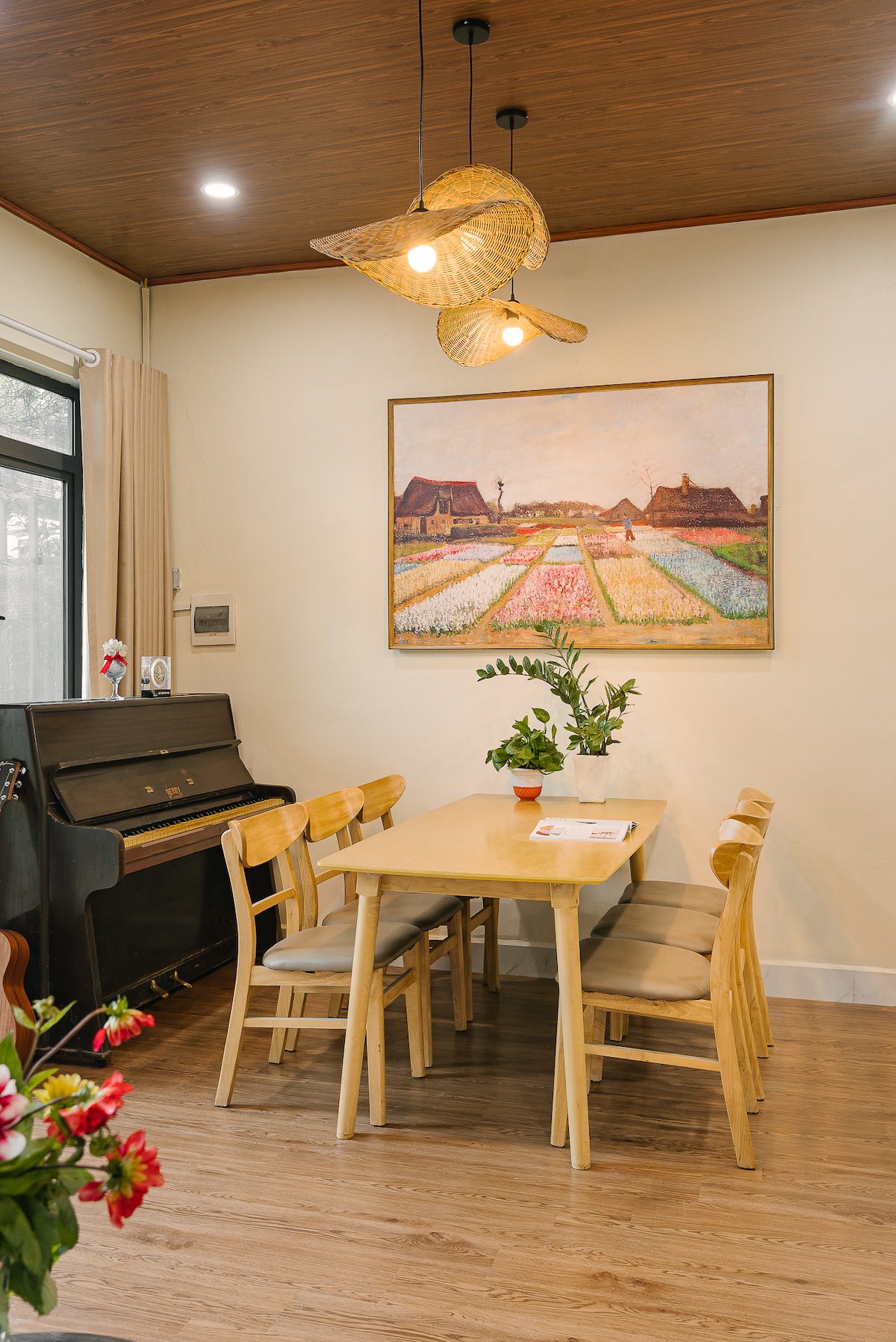 A dining area is presented with a light wooden table surrounded by six matching chairs. A potted plant decorates the center of the table. A large painting depicting a rural landscape adorns the wall above, complemented by two unique pendant lights.