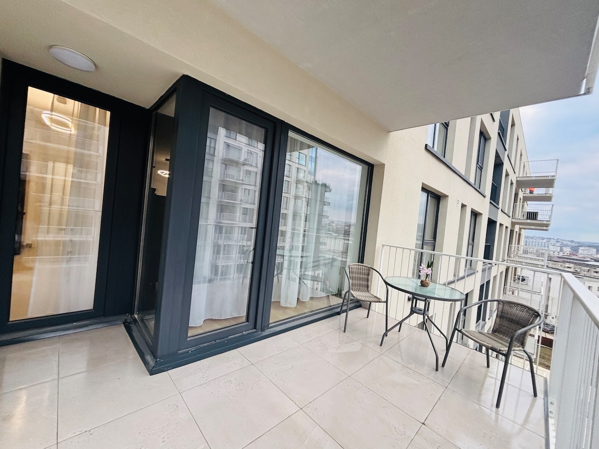 A spacious balcony is visible, featuring a glass table and two woven chairs. Large glass doors lead inside, enhancing natural light. The balcony's flooring is smooth, and the surrounding architecture is modern, creating a serene outdoor seating area.