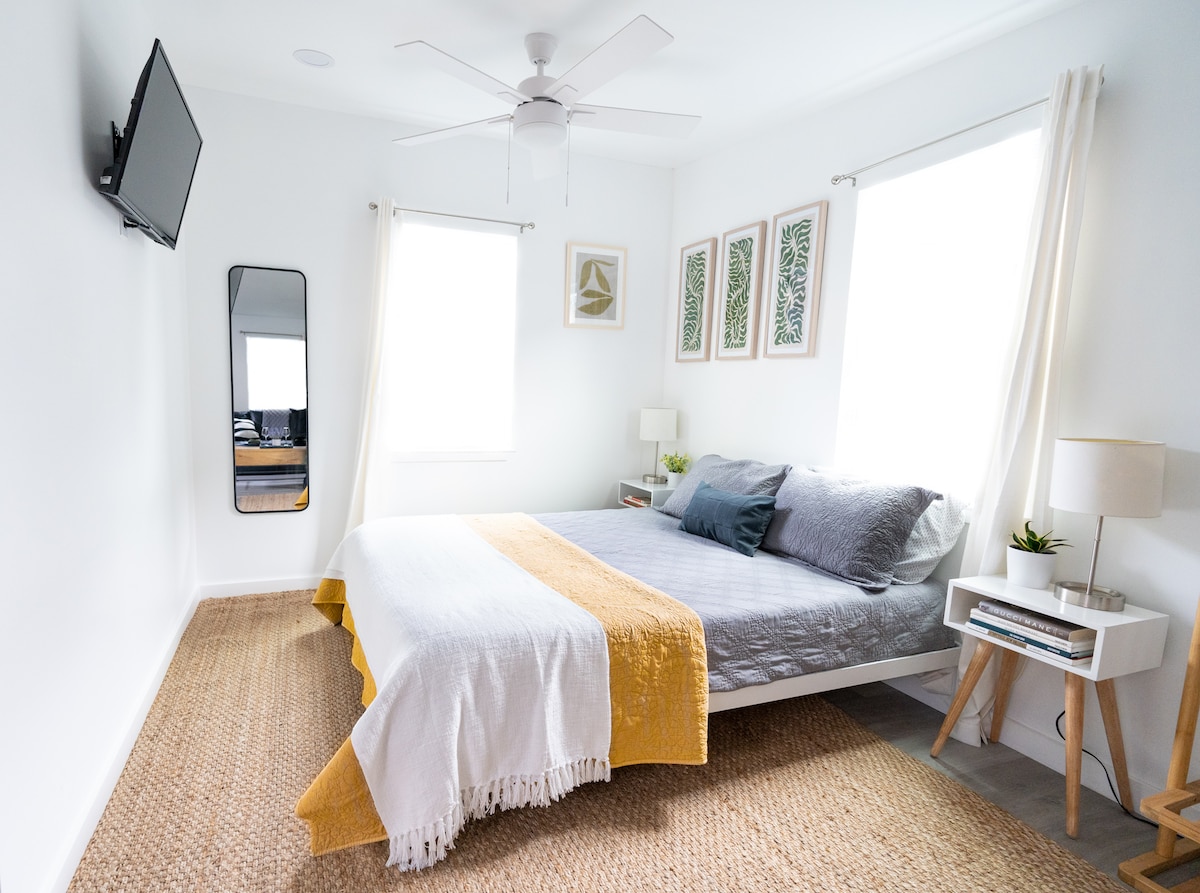 A bright and airy bedroom is featured, equipped with a spacious bed layered with a gray and yellow blanket. Light filters through two large windows, and a full-length mirror reflects the room’s layout. A lamp and small plant adorn the bedside table.