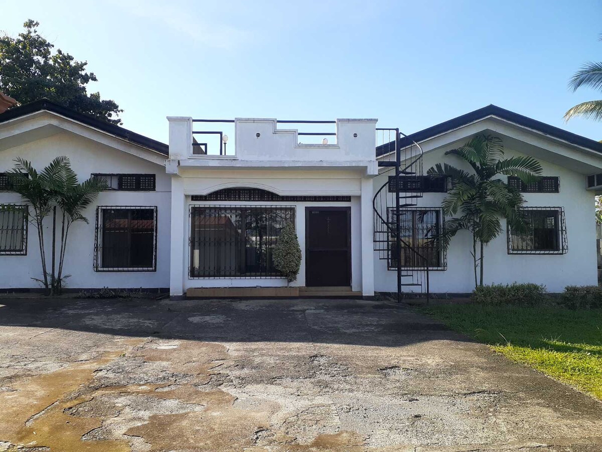 The exterior of a house is showcased, featuring a white facade with a dark roof. Two symmetrical wings extend from the central entrance, which is flanked by greenery. A spiral staircase leads to a rooftop space, surrounded by clear blue skies.