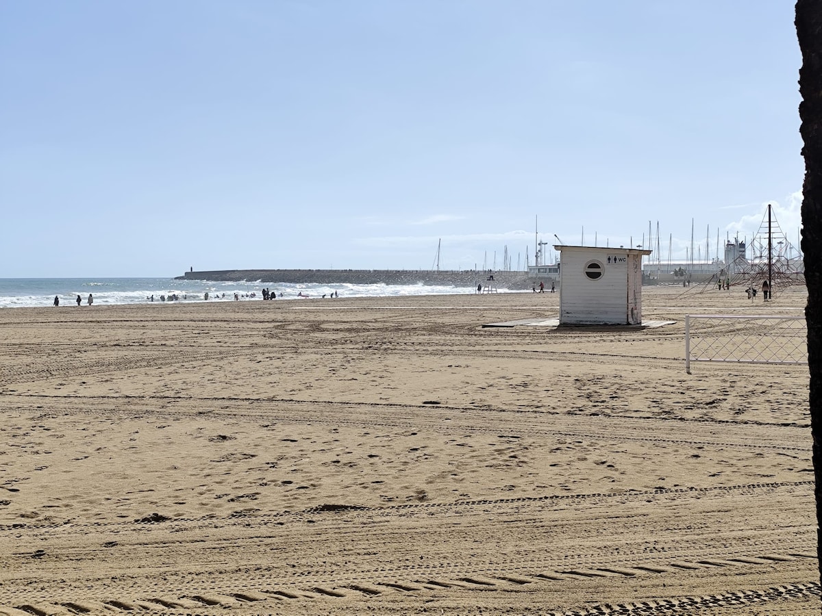 A sandy beach is visible, with gentle waves lapping against the shore. In the distance, people can be seen enjoying the water. A small beach hut stands on the sand, framed by a clear blue sky and distant sailboats along the horizon.