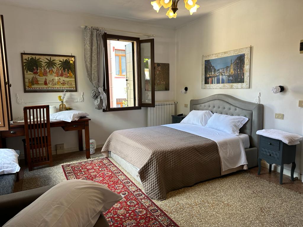 A spacious bedroom is furnished with a large bed draped in white linens. Natural light enters through two windows, complementing the decorative artwork displayed on the walls. A small desk and chair are positioned near the window, adding to the inviting atmosphere of the room.