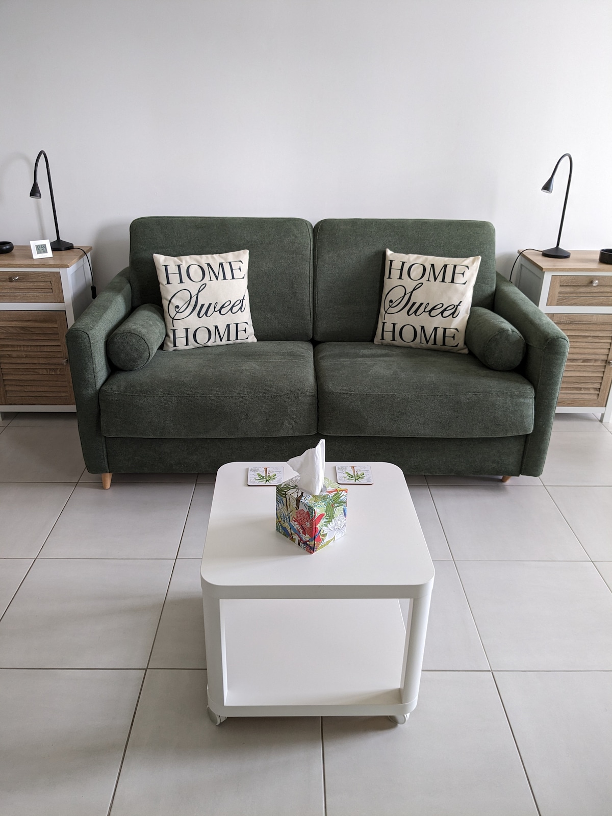 A green sofa is positioned against a plain wall, adorned with two decorative pillows that read 'HOME Sweet HOME.' A small white table sits in front of the sofa, completed by a tissue box with colorful floral designs. Two lamps provide soft lighting from either side.