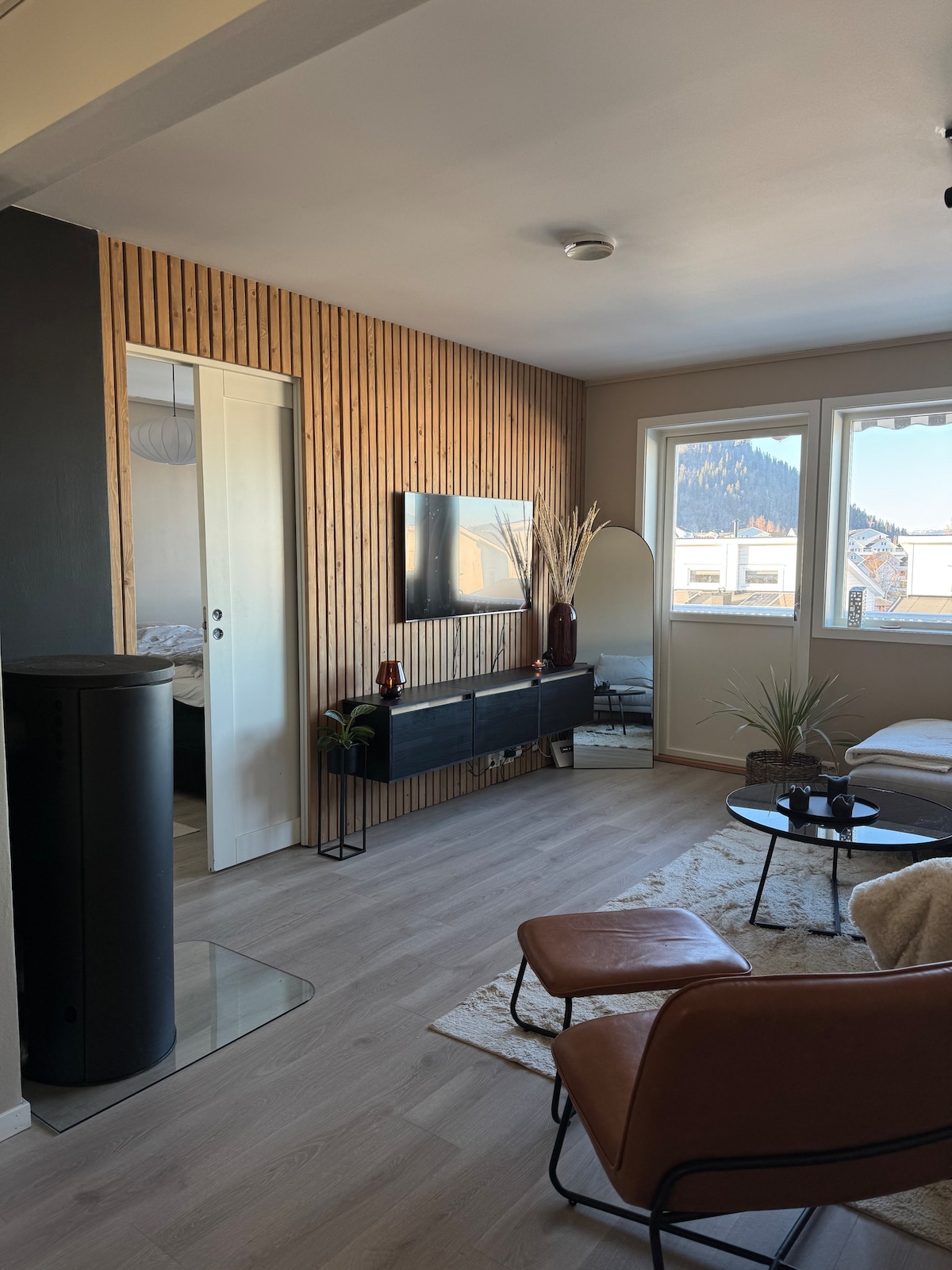 A living area is showcased with a modern aesthetic, featuring wooden accent walls and large windows that allow natural light to brighten the space. A comfortable chair is positioned beside a low coffee table, complemented by decorative plants. A television is mounted on the wall, enhancing the room's functionality.