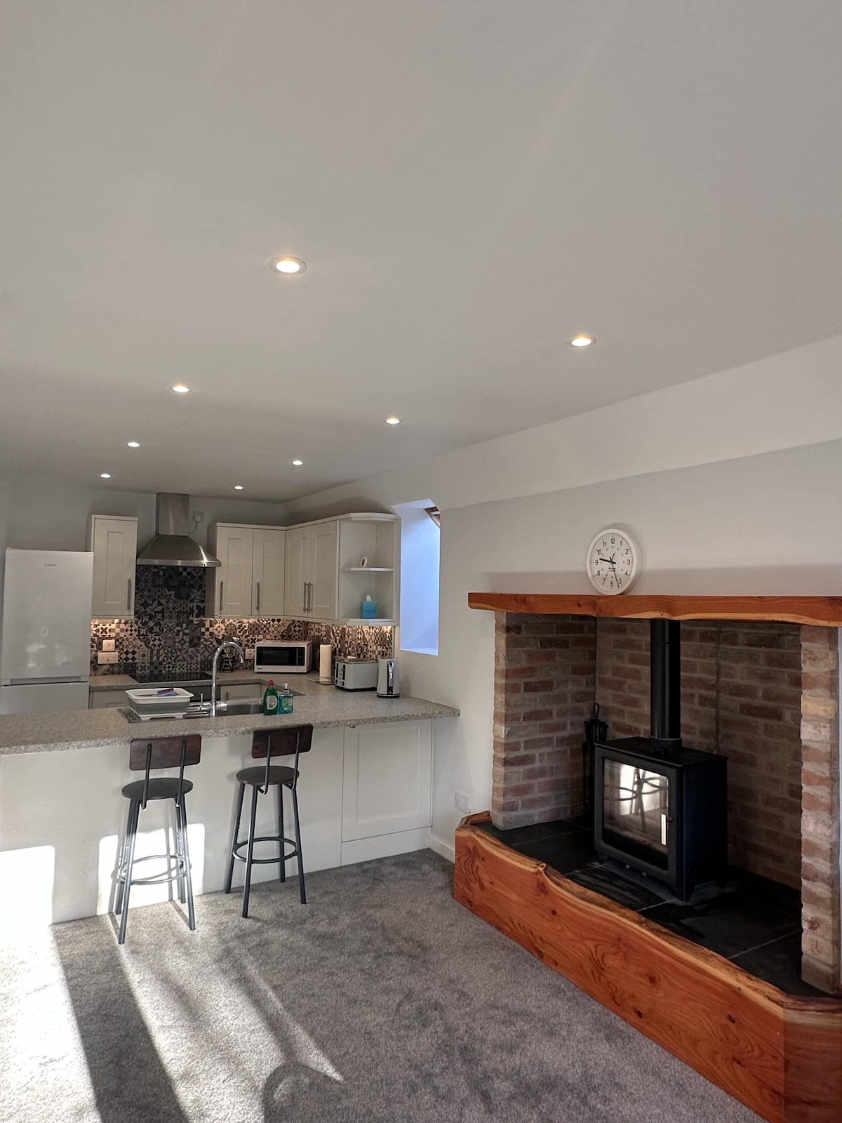 A modern kitchen presents sleek cabinetry with stainless steel appliances and a countertop, complemented by bar stools. A cozy brick fireplace adds warmth and character, while daylight enters through a nearby window, enhancing the inviting kitchen atmosphere.