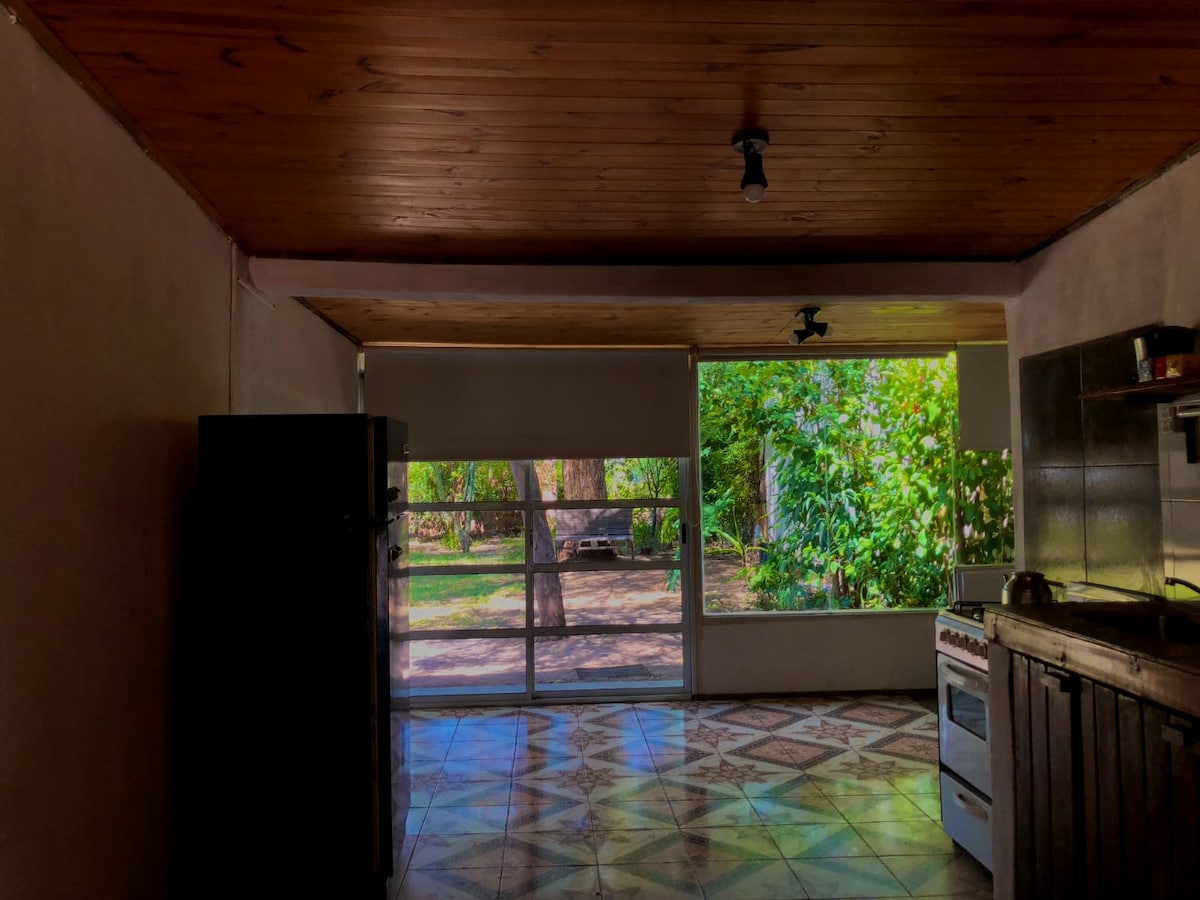 A kitchen area features a large window that offers views of a lush garden. Neutral-colored walls complement wooden ceiling beams. A refrigerator and a stove are visible, and patterned tiles cover the floor, creating an inviting space filled with natural light.