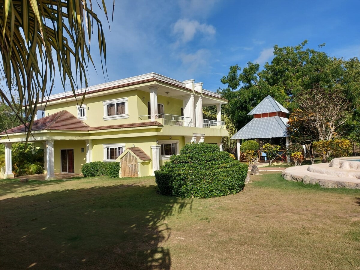 A two-story building is framed by lush greenery and a well-maintained lawn. A gazebo stands nearby, complemented by several trees. The soft hues of the exterior blend harmoniously with the natural surroundings, while the sky offers a clear, blue backdrop.