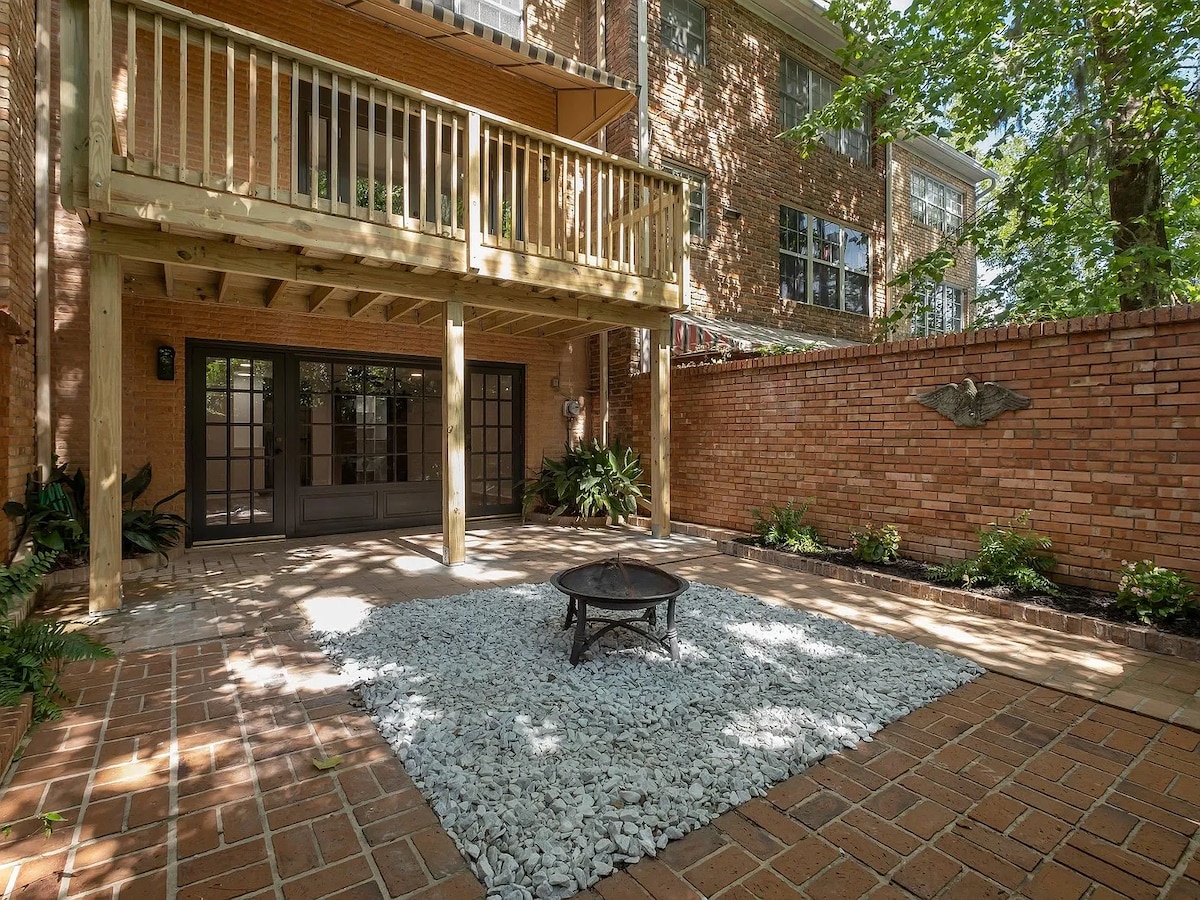 A private outdoor patio is showcased, featuring decorative gravel and pavers surrounded by lush greenery. A fire pit is centrally located, while a wooden deck above offers additional shaded space. The brick wall provides privacy, enhancing the serene ambiance of the area.