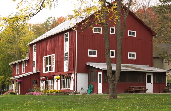 A three-story red barn is surrounded by lush greenery and seasonal flowers. Large windows enhance the barn's rustic charm, while a screened porch and picnic table invite outdoor gatherings. The well-manicured lawn offers ample space for relaxation and recreation.