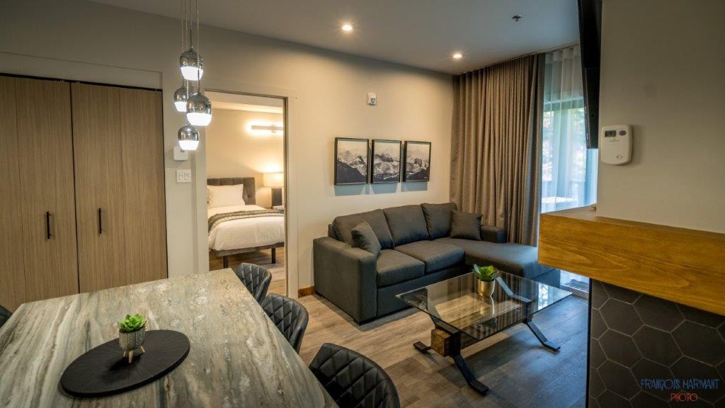 A modern living area features a comfortable gray sofa and a glass coffee table. Cozy dining space is provided with a wooden table and stylish black chairs. Light fixtures hang above, while a large window offers natural light. Two mountain-themed artworks are displayed on the wall.