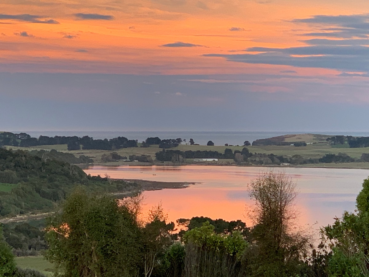 A serene landscape at sunset displays vibrant colors reflecting off the calm waters of the Catlins estuary. Lush greenery lines the foreground, while distant trees and rolling hills are visible along the horizon, enhancing the tranquil scene.