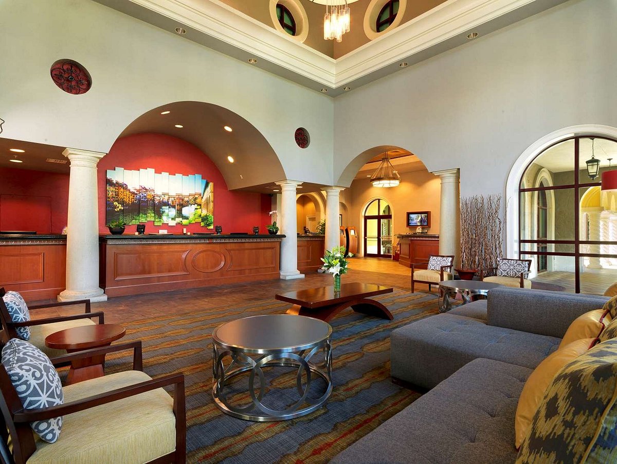 The spacious resort lobby features high ceilings and elegant arches. A warm color palette is highlighted by a red accent wall adorned with artwork. Comfortable seating arrangements include stylish chairs and a sectional sofa, complemented by a decorative coffee table and a variety of natural greenery.
