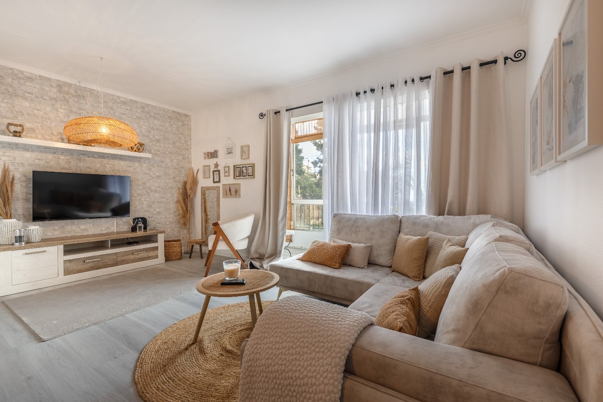 A spacious living area features a light-colored sectional sofa adorned with textured cushions. A round coffee table sits on a natural woven rug, and a television is mounted on a light wooden entertainment unit. Abundant natural light filters through sheer curtains, illuminating the space.
