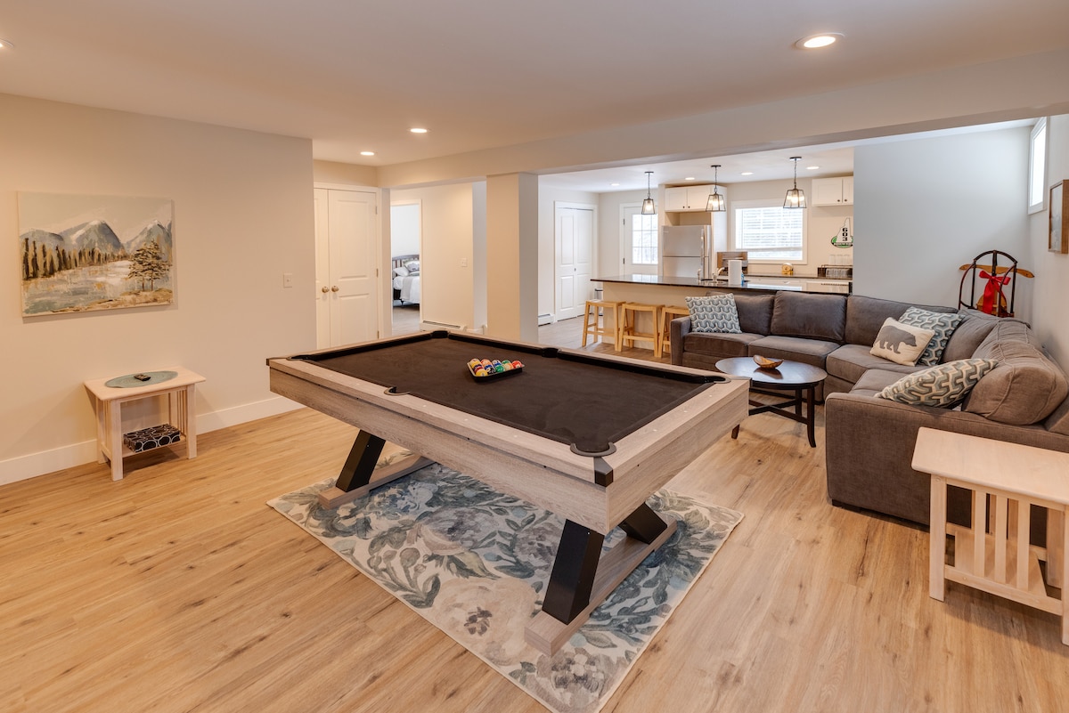 A spacious game room features a sleek pool table centered on a patterned rug. Light wood flooring extends throughout the area. A comfortable sofa and side tables are positioned against the wall, providing a relaxed seating option. A kitchen space is visible in the background.
